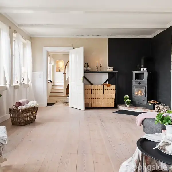 En lys stue med brændeovn i hjørnet, omgivet af trævægge og møbler. Kurv med brænde står ved siden af ovnen. Store vinduer slipper naturligt lys ind.