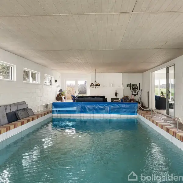 Swimmingpoolen ligger indendørs i et rum med hvide murstensvægge. Vandet er roligt, og der står møbler ved den ene ende. Vinduer langs væggen lader lys ind.