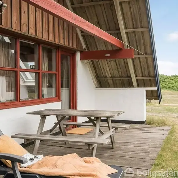 Træmøbler står på en træterrasse, herunder et bord og to stole med en pude. Hytten har store røde vinduesrammer og ses i en naturgræsindrammet område.