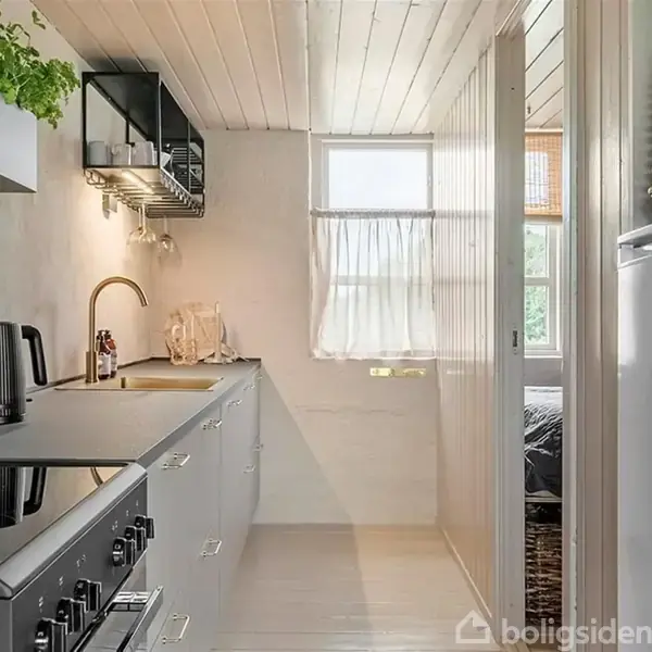 Et smalt køkken med grå skabe og en kogeplade. En elkedel står på bordet ved siden af vasken. Et vindue med gardin lyser rummet op.