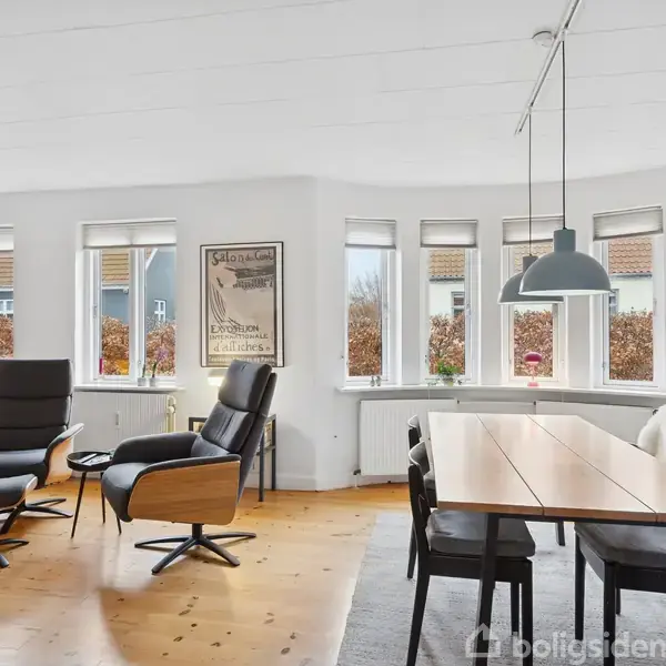 Grå hus med rødt tegltag står stille på en velplejet græsplæne omkranset af buske. Gennemsigtigt tag beskytter en lille terrasse ved siden af hovedbygningen.