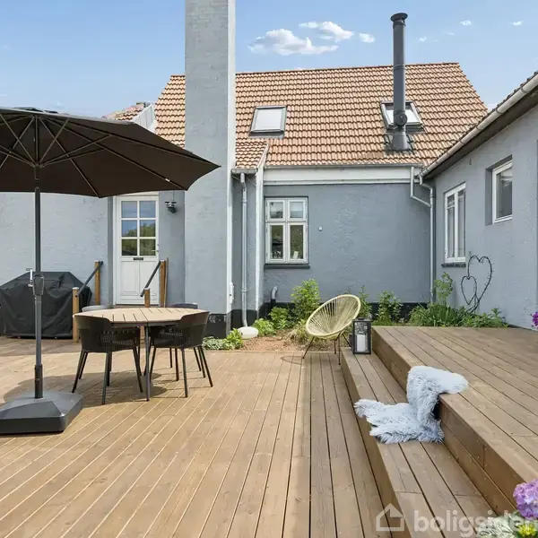 Et gråt hus med rødt tag står bag en hæk. Det har flere vinduer og en hvid hoveddør. Omgivet af andre bygninger og en blå himmel.