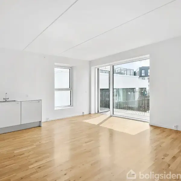 Lyst rum med trægulv og et enkelt køkkenmodul til venstre. Store vinduer og dør til altan slipper naturligt lys ind. Lejligheden ser moderne og minimalistisk ud.