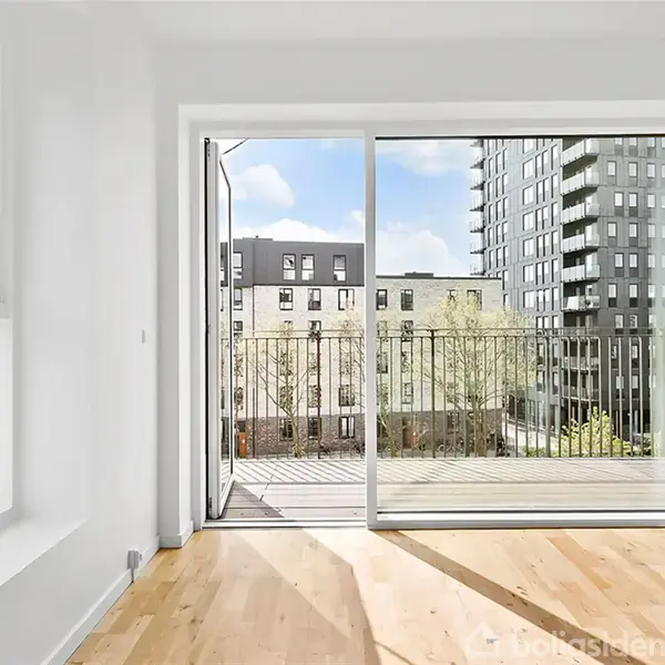 Et lyst rum med trægulv og store glasdøre åbner ud til en altan. I baggrunden ses moderne bygninger mod en blå himmel med skyer.