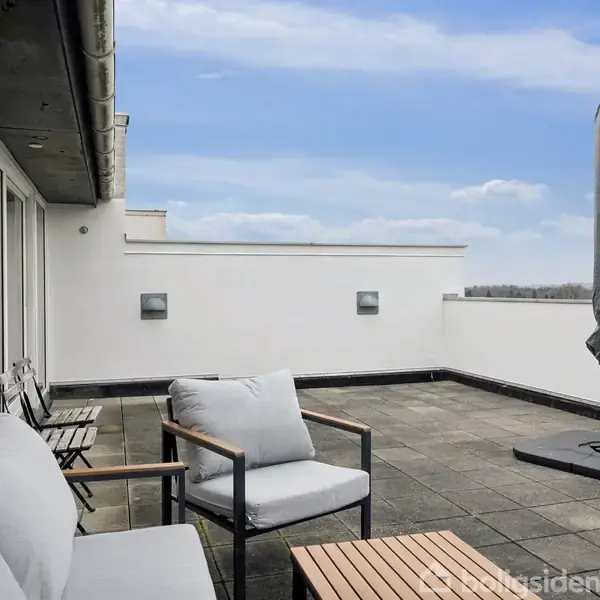 Terrassemøbler står stillet op på en rummelig tagterrasse med flisebelægning. En lukket parasol står ved siden af en sofa og stol, omgivet af en hvid mur mod en blå himmel.