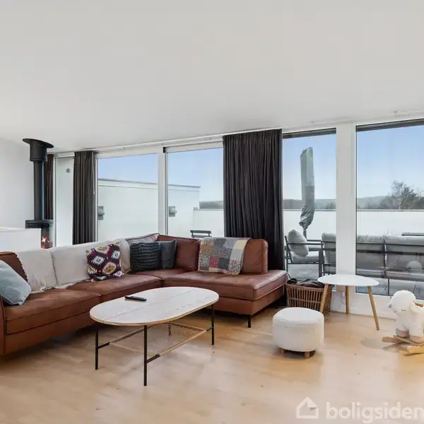 Brun L-formet sofa står i en lys stue med trægulve, store vinduer og en pejs i hjørnet. Der er en lille række borde og en legetøjshund på gulvet.