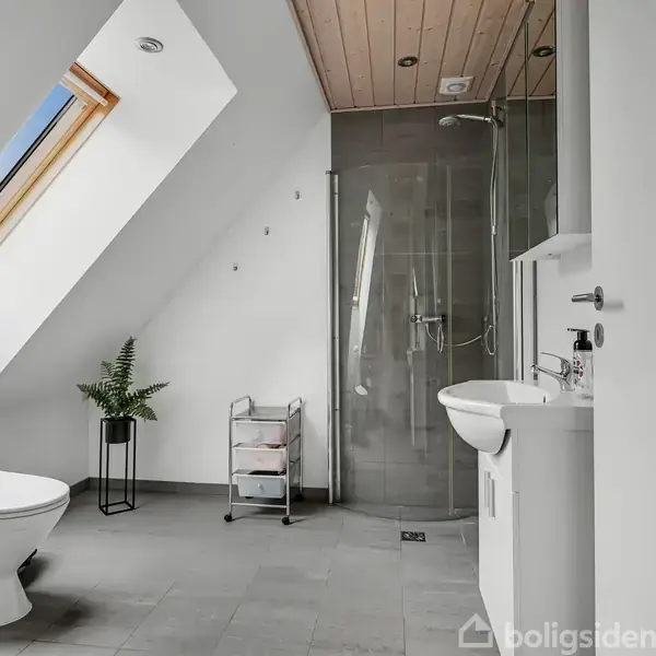 Et moderne badeværelse med et skråtag. Toilet og håndvask står tæt sammen. Et brusebad med glasvæg er i baggrunden. Et lille opbevaringsstativ og en plante er også til stede.