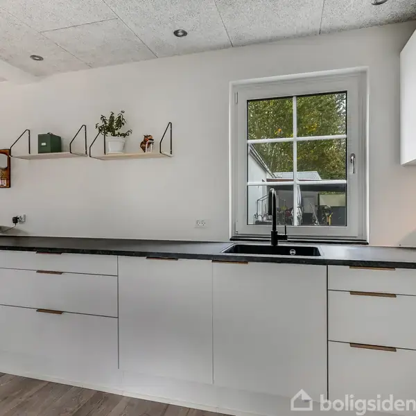 Hvidt køkken med sorte bordplader, hvor skuffer og skabe står ubrugte. Vinduet i midten åbner op til en grøn have. Hylder med planter tilføjer dekoration.