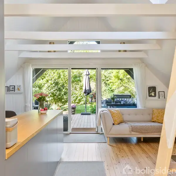 En stue med en lys sofa og et loft af træbjælker. Store glasdøre fører ud til en terrasse med en frodig have udenfor. Pejs til højre.