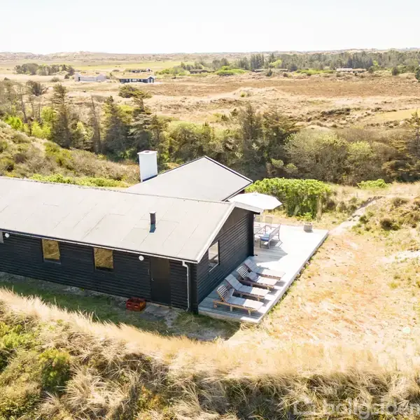 En sort, lang bygning med terrasse ligger omgivet af sandklitter og lavt bevoksede områder. Solstole står på terrassen. På afstand ses skov og åbne marklandskaber. Ingen tekst fundet.