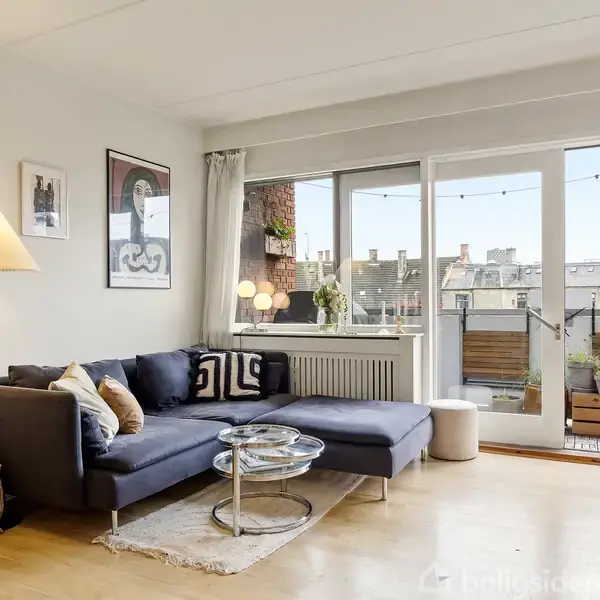 Sofa med puder står i et lyst opholdsområde nær en dør til en altan. Lampe og små borde med planter tilføjer hygge. Ingen tekst er synlig.