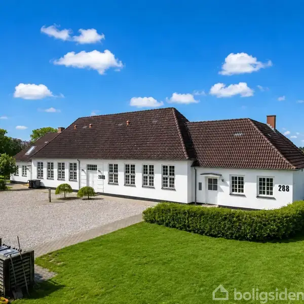 Et hvidt hus med rødt tegltag står på en grøn plæne. Numrene "288" ses på væggen. Grusvej løber langs siden, omgivet af buske og træer under en blå himmel.