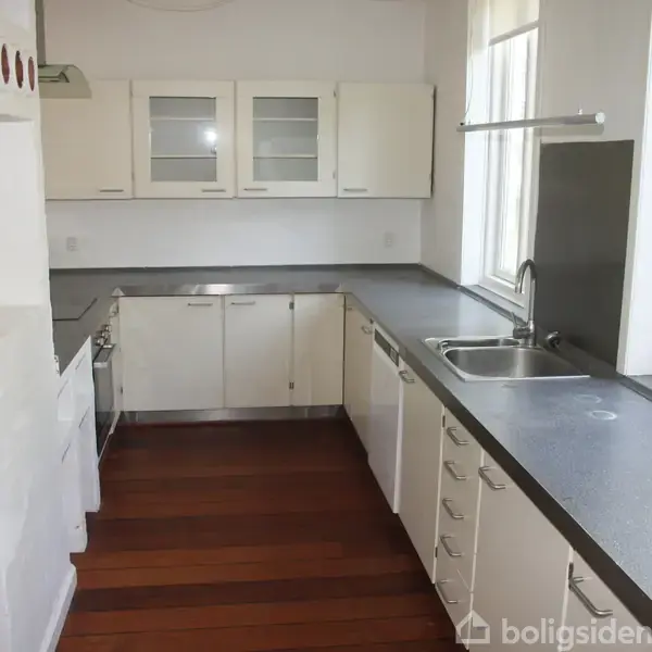 Et hvidt moderne køkken med grå bordplader, en vask, og træparketgulv. Køkkenskabe med glaslåger hænger på væggen. Store vinduer giver udsyn til en grøn have.