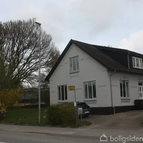 Et hvidt, to-etagers hus står stille ved en vej i et grønt område med træer. En skilt ved siden af huset viser ordet "Langgade."