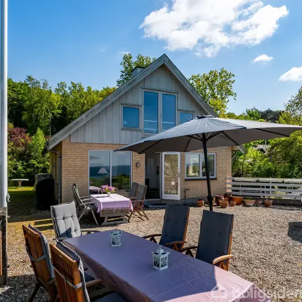 Et moderne hus med store vinduer står i en solrig have. En terrasse med siddepladser, parasol og grill omgiver det, omgivet af grønne træer og en lys blå himmel.