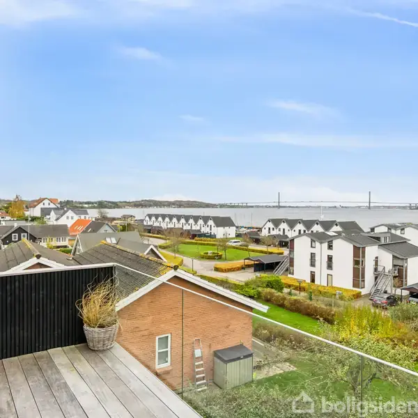 Altan med plantekrukke står stille og giver udsigt over boligområde ved vandet og broen i det fjerne. Huse spredt langs vejen, og en dansk flagstang ses i baggrunden.