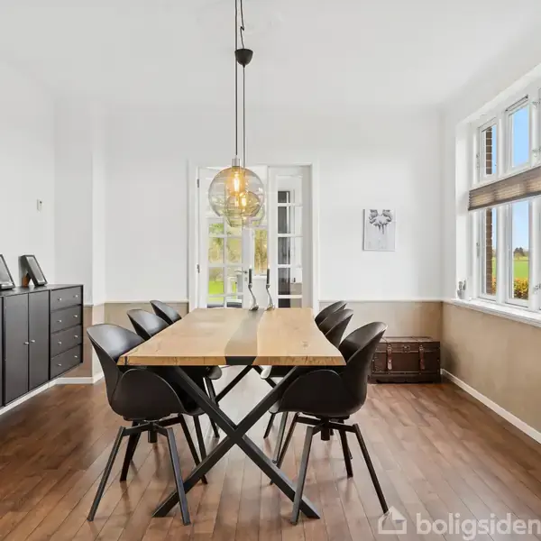 Et spisebord i lyst træ står centralt med seks sorte stole omkring. En lampe hænger over bordet. Rummet har hvide vægge, et stort vindue og mørkt trægulv.