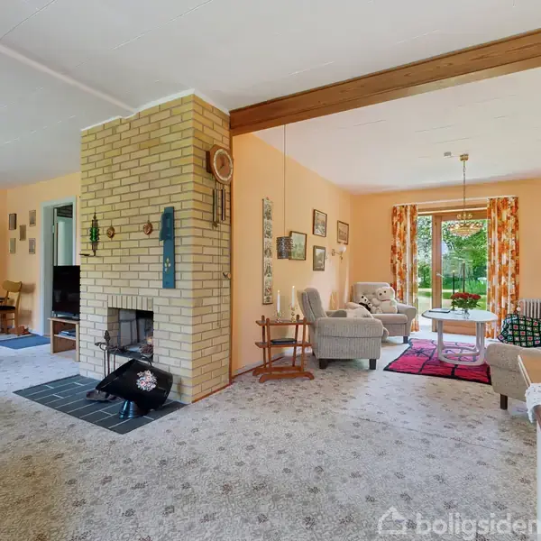 En stue med brændeovn står centralt; komfortable sofaer omgiver et sofabord. Rummet er lyst og indbydende med billeder på væggene og udsigt til en grøn have gennem store vinduer.