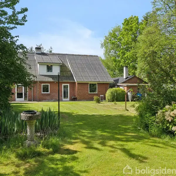 Rødt murstenshus står stille i en grøn, frodig have med tæt beplantning, omgivet af træer under en klar blå himmel. En fuglefoderstation ses i forgrunden.