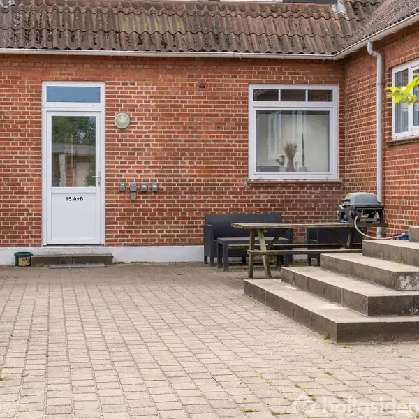 En murstensbygning med en hvid dør markeret "19A/B" til venstre. Foran er der en gårdhave med fliser, en trappe, og der står en grill og havemøbler.