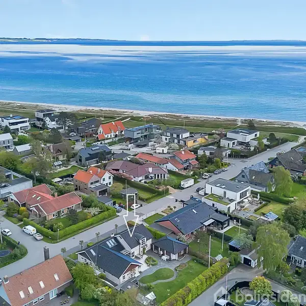 Huse ses fra luften, stille liggende på række nær stranden. Omkringliggende huse har grønne haver. Havet strækker sig i baggrunden under en blå, klar himmel. En pil peger på et specifikt hus.