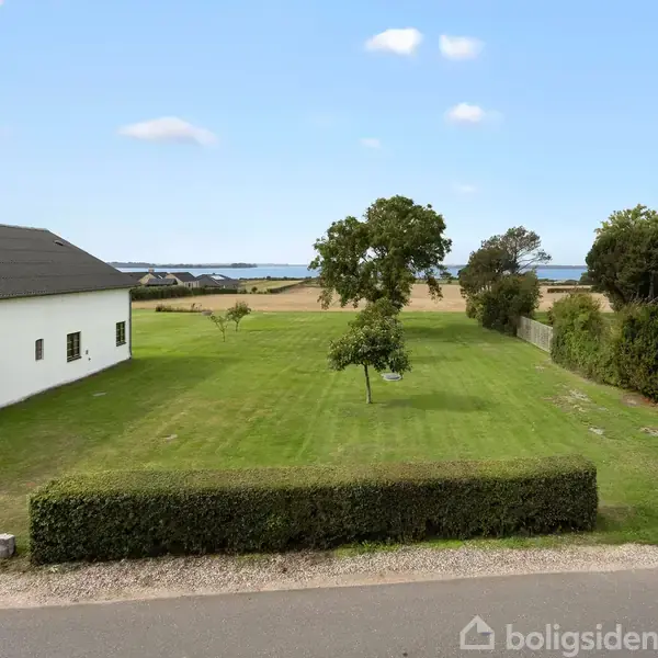 En stor græsplæne med enkelte træer ligger mellem en hvid bygning og et hus. I baggrunden ses marker og vand, under en klar blå himmel.