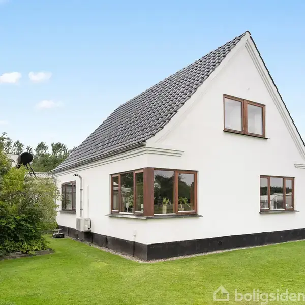 Hvidt hus med sort tag står på en velplejet græsplæne; omgivet af buske og træer, med blå himmel i baggrunden.