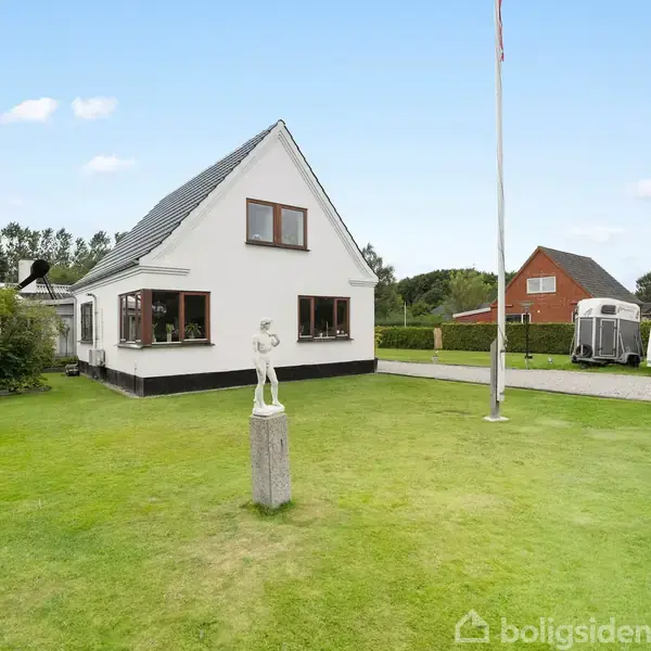 Huset står på en velplejet grøn græsplæne med en lille statue i forgrunden. En flagstang står ved siden af, og en campingvogn ses i indkørslen i baggrunden.