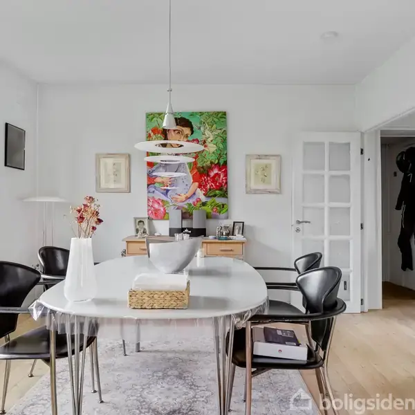 Et ovalt spisebord med sorte stole står i et lyst rum. En vase og kurv er på bordet. Der er kunst på væggen og en dør til en anden del af huset.