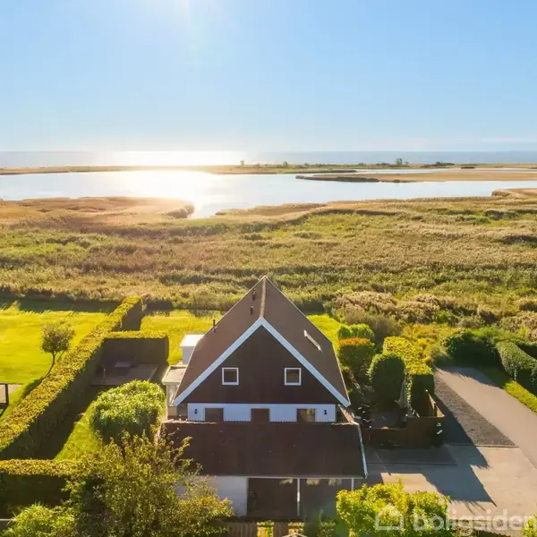Et hus ligger roligt, omgivet af grønne marker og buske. I baggrunden ses en sø, hvor solen skinner klart på vandet, og himlen er blå.