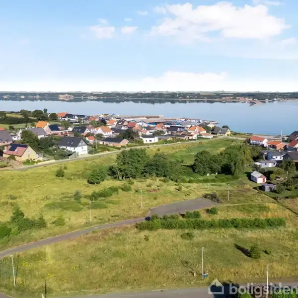 Grønt landskab med spredte huse ses foran en bred fjord, der strækker sig i horisonten, omgivet af skovområder og blå himmel.