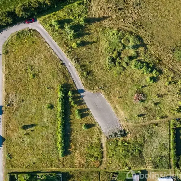 Vejen strækker sig gennem et grønt landskab med græs og spredte buske. Til venstre for vejen ses to bygninger. En rød bil er parkeret øverst til venstre.