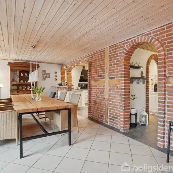 Træbord med stole står i en spisestue. Murstensvægge og buede døråbninger præger rummet. Lyse fliser på gulvet og træloft supplerer det rustikke miljø. Planter og lamper tilføjer hygge.