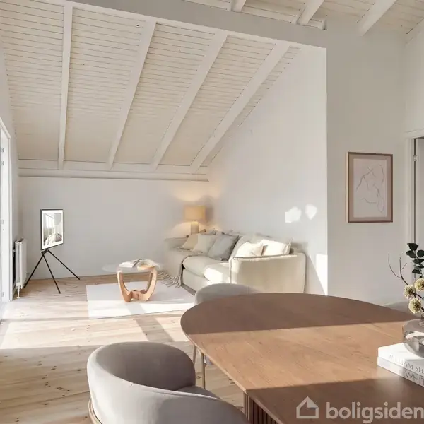 En lys stue med hvidmalede trævægge og skrå loftbjælker. En grå sofa står mod væggen, oplyst af en bordlampe. Et rundt bord og stole står i forgrunden. En plante og bøger er placeret på bordet.