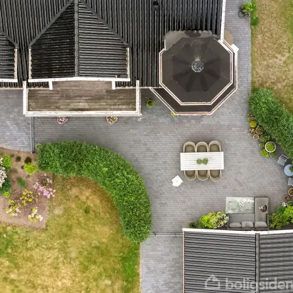 Hus med sort tag set oppefra, omgivet af terrasse med spisebord og stole. Omkring terrasse er der grønne buske og blomsterbede med farverige blomster. Græsplæne omgiver området.