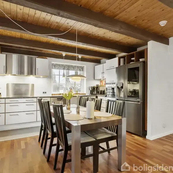 Et spisebord med seks stole står i et køkken med træloft, synlige bjælker og hvide skabe. Et køleskab og en ovn ved siden af. Vinduer giver naturligt lys.