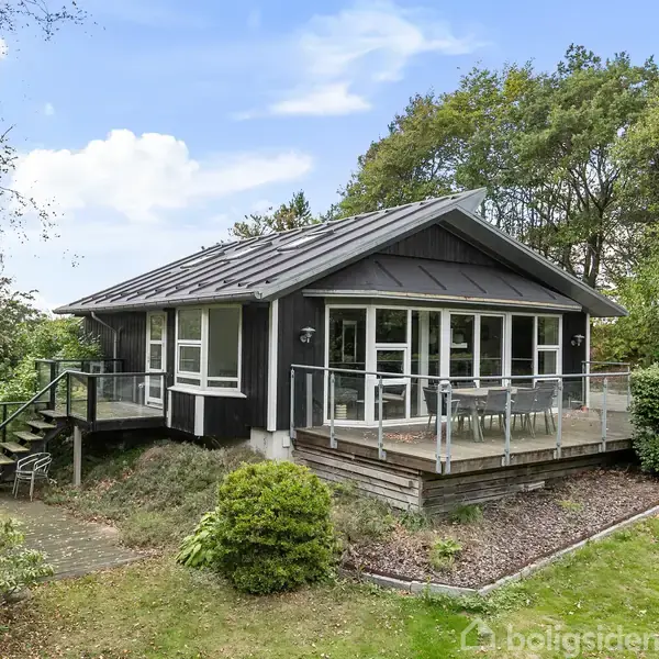 Et sortmalet sommerhus med en træterrasse omkranset af glasrækværk. Terrassen har havemøbler, og huset er omgivet af græsplæne og træer, der skaber en naturskøn atmosfære.