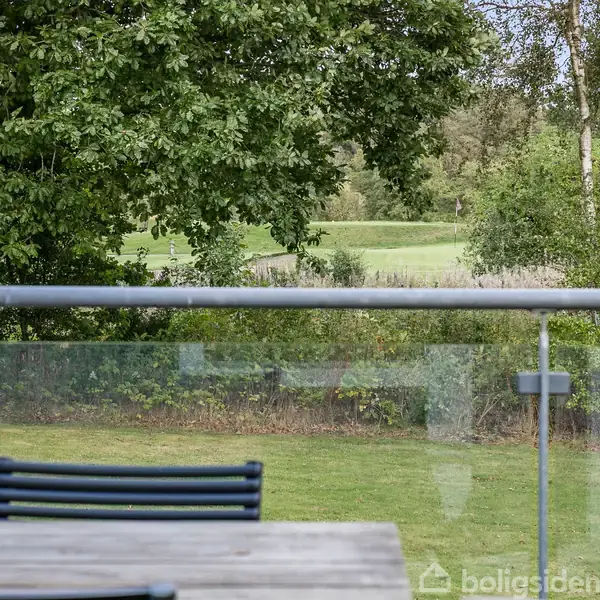 En bordplade med stole omkring står på en terrasse med glasrækværk. I baggrunden ses en græsplæne og grønne træer.