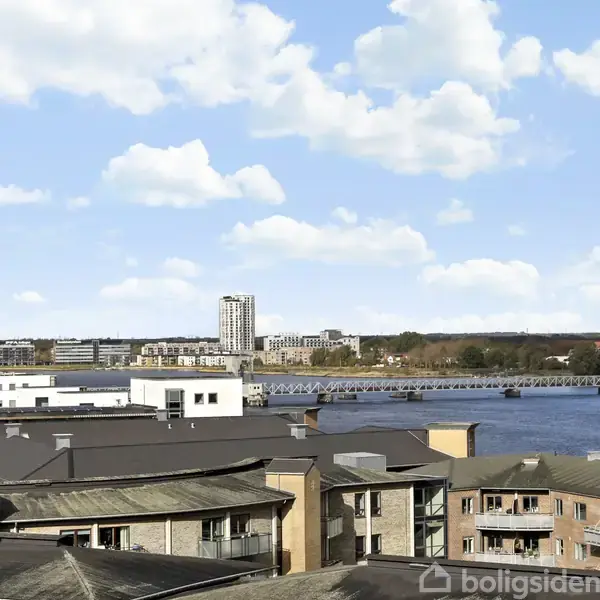 Bygninger står stille ved en flod med en bro i baggrunden, omgivet af flere moderne lejlighedskomplekser. Klar blå himmel med lette skyer skaber en lys og rolig stemning.