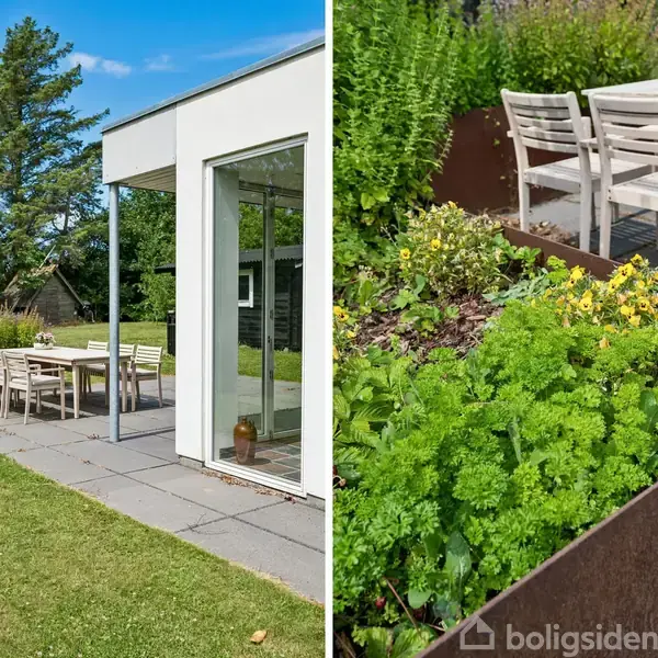 Hvidt hus med udendørs terrasse i en have. På terrassen står et bord med stole omgivet af grønne planter i højbede. Solrig dag med blå himmel.