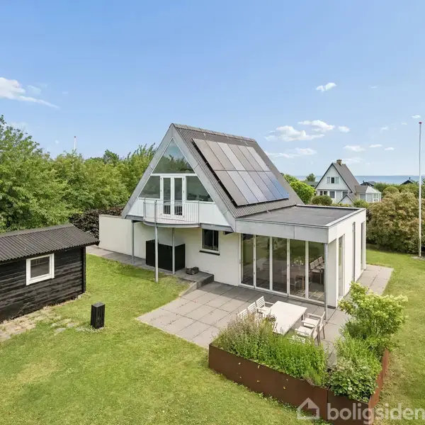Et moderne A-formet hus med solpaneler på taget står omgivet af en velplejet have. Et fritliggende sort skur er placeret til venstre. Himlen er klar med få skyer.
