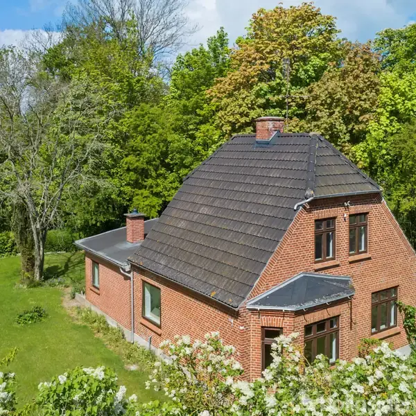 Rødt murstenshus står i en frodig have med træer og buske. Taget er mørkt og har en skorsten. Himlen er blå med spredte skyer.