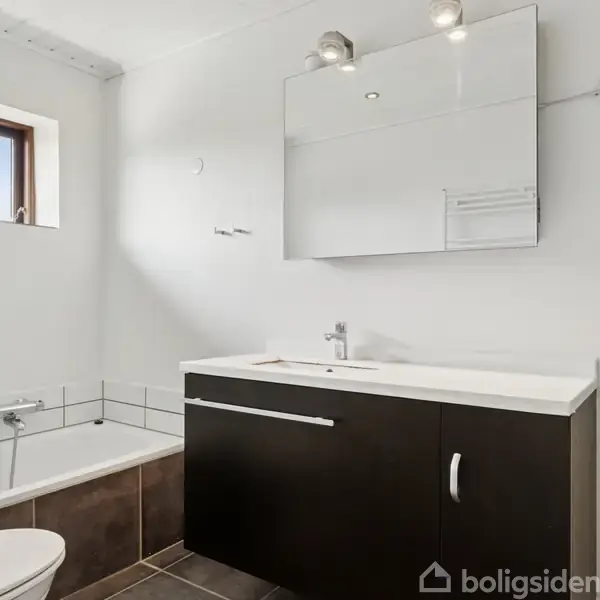 Et badeværelse med et mørkt skab under en hvid vask, spejlskab med lys ovenover, og et badekar ved siden af. Et vindue over badekarret giver naturligt lysindfald.