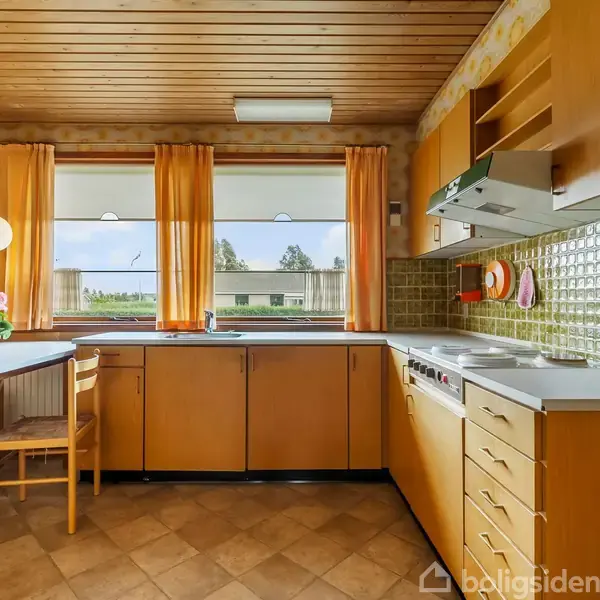 Køkken med retro træskabe og grønne fliser, bord og stole ved siden af. Vinduet viser en udsigt med træer, og køkkenet har gule gardiner og en pendellampe.