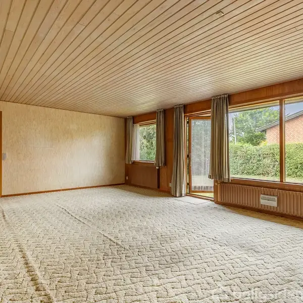 Værelse med lyse tæpper på gulvet og træpaneler i loftet. Store vinduer med gardiner giver udsigt til en have. Dør til venstre fører til en anden del af huset.