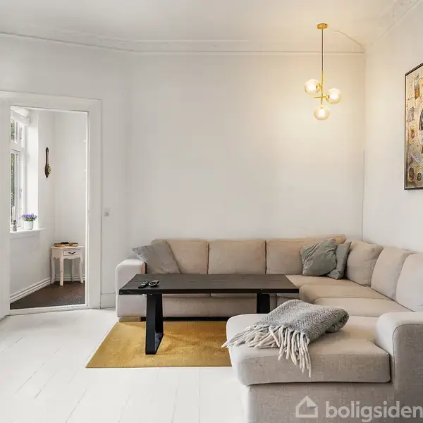 En beige hjørnesofa står i en lys stue med hvide vægge. En sort sofabord står foran sofaen. Der hænger en moderne loftslampe og kunst på væggen. En dør fører til et andet rum.