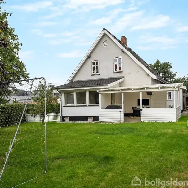 Et hvidt hus med en overdækket terrasse står i en grøn have med et fodboldmål og en oppustelig pool. Træer og buske omkranser området.