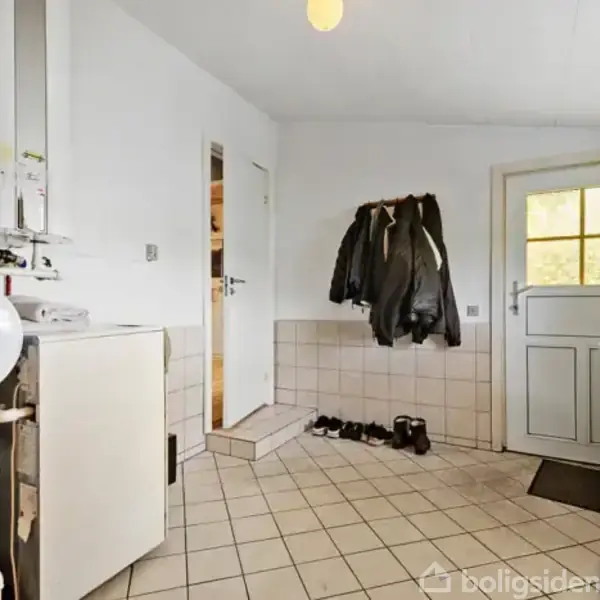 Væghængt kedel står i funktionel entré med jakker hængende på knage, sko på gulvet og dør til højre. Fliser på gulv og under væg.