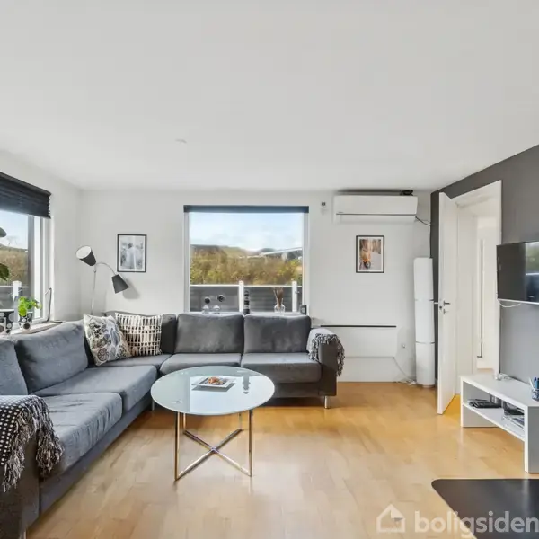 En stue med en stor grå hjørnesofa står ved siden af et rundt glasbord. En brændeovn er til højre, og væggene har hængende billeder. Lyset strømmer ind gennem vinduerne.