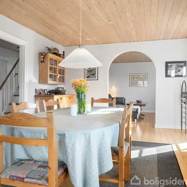 Et rundt spisebord med en lyseblå dug og blomster på står i et lysfyldt rum med trægulv og hvide vægge. En lampe hænger over bordet.